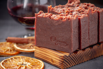 Pieces of handmade natural wine soap on a wooden soap dish, a glass of wine and pieces of orange on a grey background