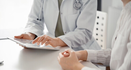 Doctor and patient sitting at the desk in clinic office. The focus is on female physician's hands using tablet computer, close up. Perfect medical service and medicine concept