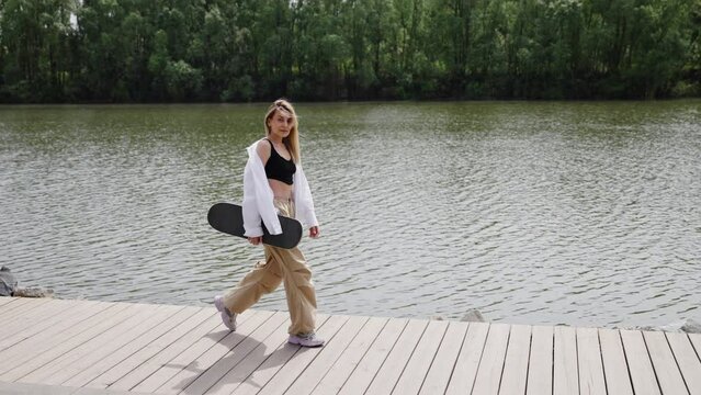 The girl walks along the embankment against the backdrop of water and a green forest. Skateboard in the hands of a beautiful girl with blonde hair. Beautiful city promenade with stones and water