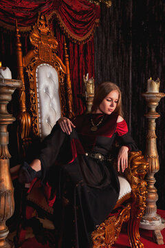 Medieval queen in an old style posing on golden throne fortress, pensive look. Bored lady in historical image sitting on antique gold throne in castle room. Concept of theatre perform. Copy text space