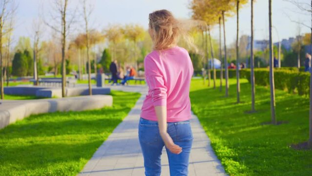 young beauty happy woman walking dancing in street modern city blonde hair fly in wind. Sexy girl smiling face. public park nature green grass trees sun light. pink shirt blue jeans casual clothing