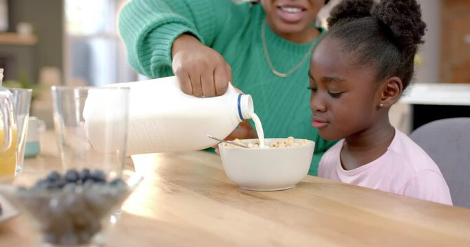 Happy african american mother pouring milk on cereal with daughter in kitchen, slow motion