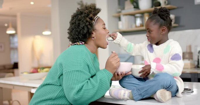 African american mother and daughter eating cereal with milk in kitchen, slow motion