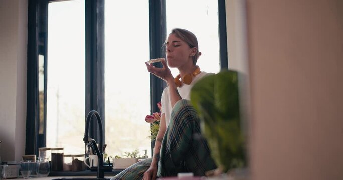 A blonde girl in a white T-shirt sits on the windowsill in the kitchen and eats a sandwich