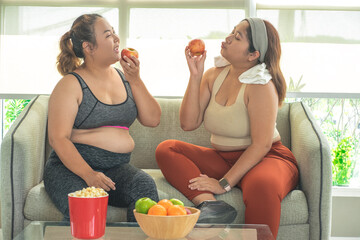 Two plus size women in sports bras sitting on the couch holding an apple eating quality food and discussing diet and exercise in the gym.