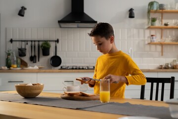 Fototapeta na wymiar A boy in a yellow sweater makes himself a sandwich at the kitchen table. The young man got hungry and decided to make himself a blueberry jam sandwich.