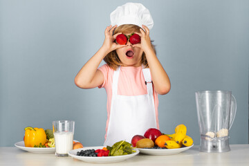 Portrait of a 7, 8 years old child in cook cap and apron hold strawberries making fruit salad and cooking food in kitchen. Cute little blonde happy smiling chef.
