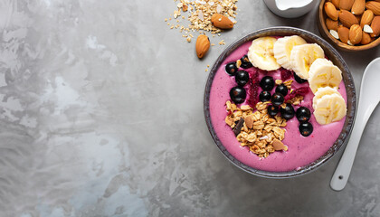 Pink black currant berries, banana smoothie bowl with granola, flakes from almonds and chocolate on light gray concrete background. Selective focus. Top view. Copy space.