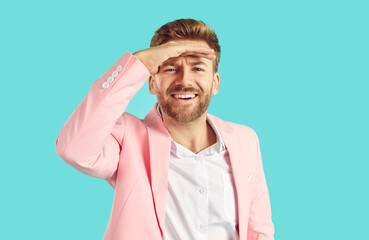 Handsome bearded young man in white shirt and pink suit isolated on blue background holds his hand on forehead like visor to make some shade above eyes and looks in distance with happy face expression - obrazy, fototapety, plakaty