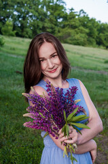 Portrait of a beautiful happy brunette girl in a blue dress in a field with a bouquet of flowers from lupines