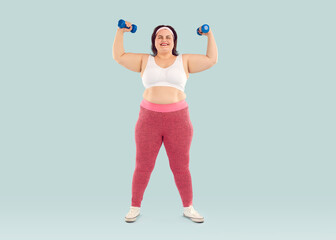 Happy chubby overweight young woman exercising with dumbbells. Full length shot of plus size, fat woman wearing sports bra and leggings doing physical exercise isolated on light blue studio background