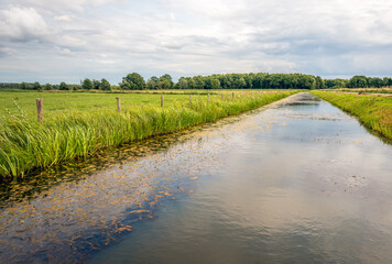 Fototapeta na wymiar Wide ditch in a Dutch polder landscape. The clouds are reflected in the smooth water surface. Along the bank grows and behind the fence of wooden posts with wires is a meadow.