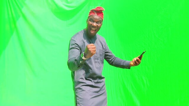 Standing Yoruba cultural man in blue traditional dress and orange cap celebrate good business news on phone and shows phone to camera