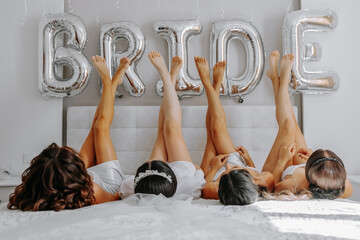 Young bridesmaids in white silk dresses on a bed in the bride's room. Beautiful women celebrating bachelorette party sitting on bed. - obrazy, fototapety, plakaty
