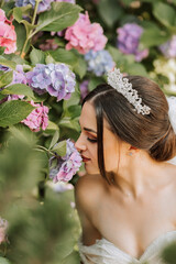 Obraz na płótnie Canvas young beautiful bride in wedding dress with open shoulders and crown on her head sitting near hydrangea flowers, fashion photo taken under harsh sunlight