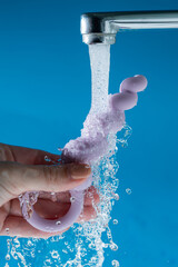 Woman holding lilac anal beads under running water on blue background. Sex toy hygiene concept.