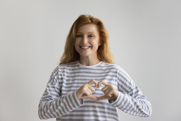 Happy naturally red haired young woman showing finger heart, making hand gesture at chest, promoting love, romance, happiness, care, kindness. Female portrait with white background