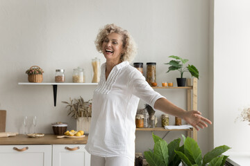 Cheerful pretty mature woman having fun at home kitchen, dancing with open arms, laughing, enjoying music, active leisure time, feeling happy, carefree, inspired