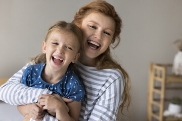 Cheerful red haired mom cuddling, tickling cute kid on sofa, playing with girl, holding shouting...