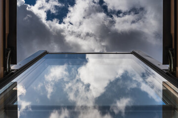 Cloudy sky frames open attic window, with ethereal clouds reflected in glass. Moody ambiance captures nature's beauty
