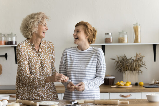 Joyful excited mom and adult daughter baking sweet homemade pastry snacks, cookies, biscuits, shaping, rolling dough, talking, discussing family recipe, smiling, laughing, having fun