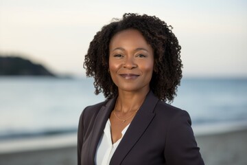 Portrait photography of a satisfied Nigerian black woman in her 40s wearing a classic blazer against a beach background 
