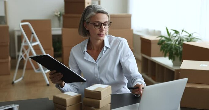 Happy grey haired small business owner woman processing order, checking boxes for sending, shipping, writing notes, keeping records, enjoying work at laptop, workplace table
