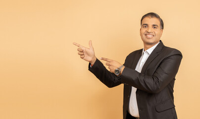 Happy mature indian businessman wearing black suit pointing at blank space isolated over beige background. Look here, Advertisement and promotion.