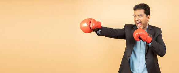 Angry young indian employee wearing boxing gloves punching isolated over beige background,...