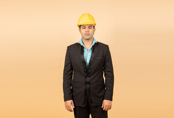 Young indian engineer or architect wearing black formal suit and yellow hard hat or helmet standing isolated over beige background. Industrial and construction concept. Copy space.