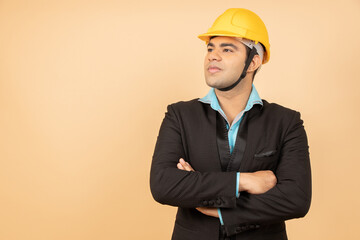 Young indian engineer or architect wearing black formal suit and yellow hard hat or helmet standing cross arms isolated over beige background. Industrial and construction concept. Copy space.