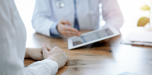 Doctor and patient sitting at the wooden desk in clinic. Female physician's hands pointing into laptop computer monitor, close up. Medicine concept