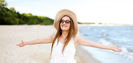 Fototapeta na wymiar Happy smiling woman in free happiness bliss on ocean beach standing with a hat, sunglasses, and open hands. Portrait of a multicultural female model in white summer dress enjoying nature during travel