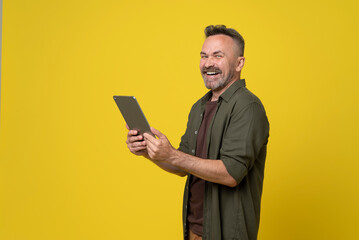 man 50s dressed in green shirt holds digital tablet pc gadget in hands and happy smiled to camera isolated on yellow background