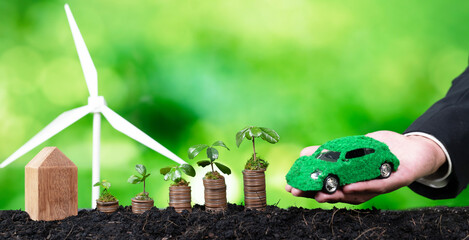 Businessman holding EV car model with seedling grow on coin stack and wind turbine. Eco-friendly...