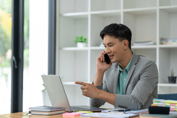 Naklejka premium Happy young asian businessman looking at data on laptop, chatting on phone using smartphone for communication. Check through the application Go online while at work in the office.