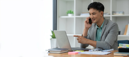 Fototapeta premium Happy young asian businessman looking at data on laptop, chatting on phone using smartphone for communication. Check through the application Go online while at work in the office.