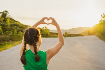 Back view girl in green T-shirt holds her hands in shape of heart against background of sun in...