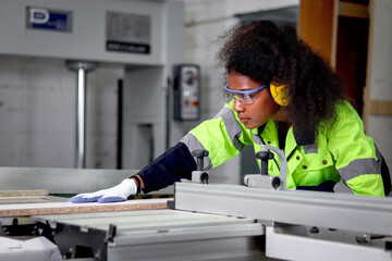 African industrial woman worker with curly hair wears safety vest and protective headphones, works with wood cutting machine, female engineer works at CNC woodworking manufacturing furniture factory.