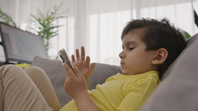 Sweet child sitting on sofa concentrated on electronic application on cellphone