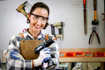 Portrait of happy smiling beautiful carpenter woman wearing safety glasses goggles and apron holding hand drill, female craft worker standing in woodworking furniture workshop, woman in carpentry shop