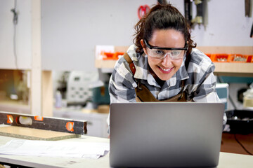 Happy smiling beautiful carpenter woman wearing safety glasses goggles and apron working on laptop computer, female craft worker with wooden furniture background in woodworking carpentry workshop.