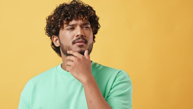 Portrait of a handsome curly-haired Indian man in a turquoise t-shirt scratching his chin thoughtfully and looking up. The emotion of thoughtfulness, doubt. The camera moves slowly on the slider.