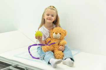 Little child girl plays with plush toy at doctor appointment. Child listens to soft toy with stethoscope. Doctors concept for children, copy space for text.