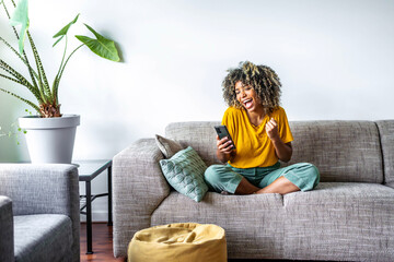 Fototapeta Excited happy young black woman holding smart phone device sitting on sofa at home - Happy satisfied female looking at mobile smartphone screen gesturing yes with clenched fist - Technology concept obraz
