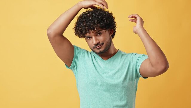 Video portrait of a handsome curly-haired young stylish Indian man in a turquoise t-shirt fixing his curly hair and looking at the camera as a mirror while standing in a studio on a yellow background.