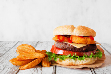 Delicious onion ring hamburger with potato wedges. Side view table scene against a white wood background.