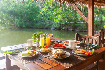 Breakfast table at Asian resort overlooking lake. Pancakes, sandwiches, coffee, juices, salad, fresh fruits. Healthy, delicious dishes on a sunny Thailand vacation.