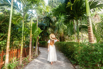 Happy tourist in white dress and straw hat strolls through a beautiful tropical forest in Thailand. Concept of nature and travel.