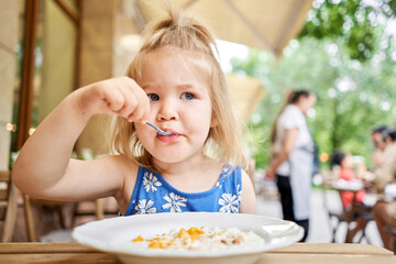 Little kid having breakfast at cafe. Adorable girl drinking still water, eating rice porridge with mango. enjoying breakfast. 